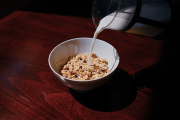 Pouring the milk into oatmeal in a white porcelain bowl on the wooden background.