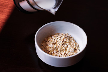 Pouring the milk into oatmeal in a white porcelain bowl on the wooden background.