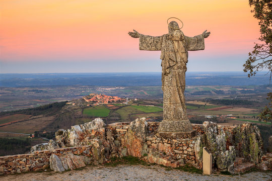 View of the Serra da Marofa viewpoint, in Portugal, at sunset, with Cristo Rei in the foreground and the historic village of Castelo Rodrigo in the background.