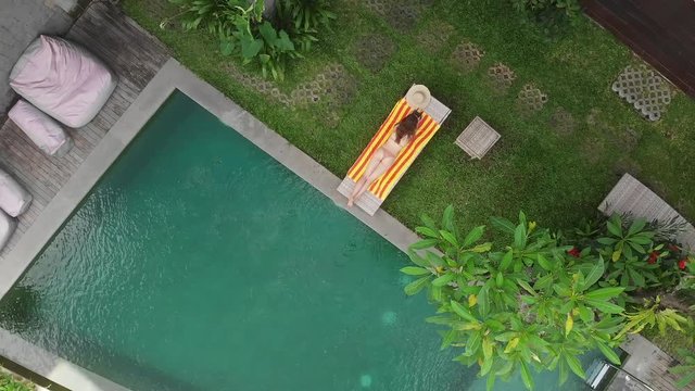 Aerial top view of slim young woman in beige bikini relaxing and get sunbathe near swimming pool in chaise-longue jungle in Ubud. Drone going up. Vacation concept.