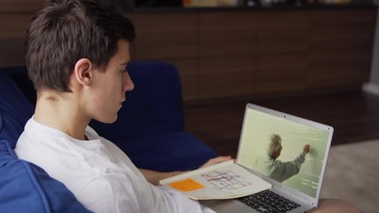 Tilt up side view of male college student sitting on sofa at home and studying online watching video lesson with teacher