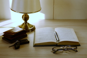 There is a table lamp on the table, an open book, a pen, glasses, a wallet and car keys.