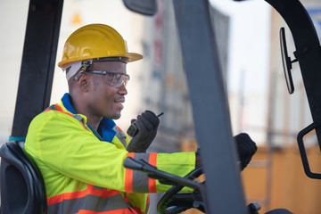 Male worker driving forklift and using walkie-talkie in shipping yard industrial container...