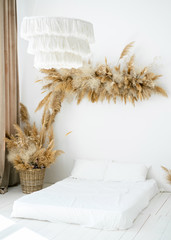 Scandinavian minimalist interior. Dry gras and pampas grass floral composition on the wall. The mattress covered with white bed linen on the floor. Stylish bedroom from the interior magazine.