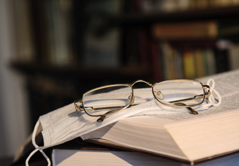 On the opened book lies a medical mask and glasses in a gold frame. Against the background of bookshelves. Concept - training, self-isolation