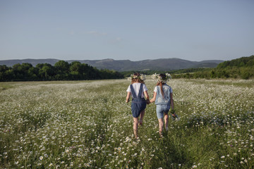 Fototapeta na wymiar two cute teen girls in denim overalls walk in a daisy field