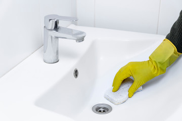 Woman is cleaning a sink in the bathroom, using clean water using yellow gloves and a white sponge.