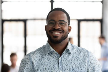 Headshot portrait of smiling African American male employee in glasses look at camera posing at workplace, happy motivated biracial man worker show confidence and leadership, employment concept