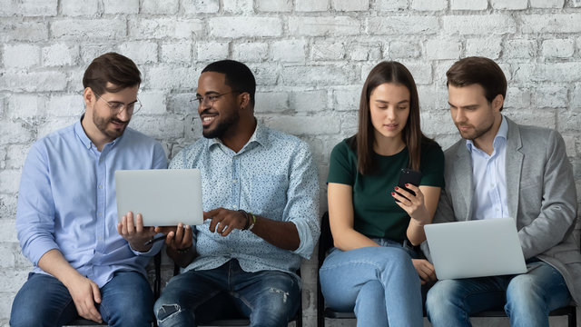 Focused diverse young people sit in queue discuss ideas work on laptops together, concentrated multiethnic job candidates prepare wait for interview in office using gadgets, employment concept