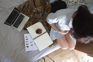 A young woman sitting on the bed looking at the pages of a notebook. Woman having a fruit bowl and a laptop on the bed for breakfast