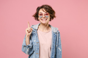 Excited young brunette woman girl in casual denim jacket, eyeglasses posing isolated on pastel pink background. People lifestyle concept. Mock up copy space. Hold index finger up with great new idea.