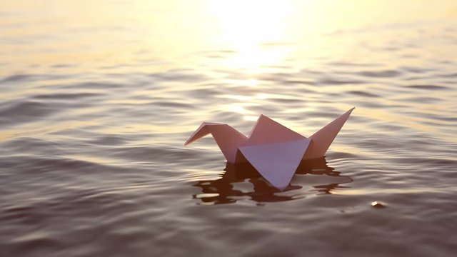 A paper crane floats through the waves in the water at a beautiful sunset. Origami crane saving lives. The concept of a dream, future, childhood, freedom or hope. Slow motion. Sasaki Origami Sadako