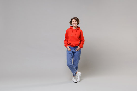 Cheerful young brunette woman girl in casual red hoodie, blue jeans posing isolated on grey background studio portrait. People emotions lifestyle concept. Mock up copy space. Holding hands in pockets.