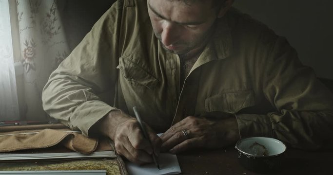 Soviet Army soldier analyzes map and draws up action plan sitting in abandoned house at table near window.