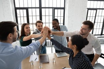 Overjoyed diverse businesspeople give high five celebrate shared victory or win at meeting, excited multiracial colleagues feel motivated engaged in teambuilding activity at briefing in office