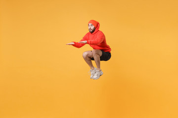 Young bearded fitness sporty guy 20s sportsman in hat, hoodie, shorts spend weekend in home gym isolated on yellow wall background. Workout sport motivation lifestyle concept. Jumping doing exercises.