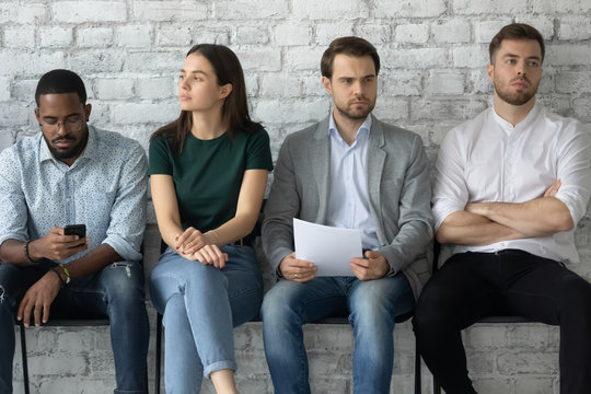 Bored young multiethnic candidates sit on chairs in line queue wait for interview in office, concentrated thoughtful diverse multiracial applicants worried before recruitment talk, employment concept