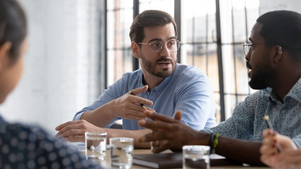 Focused multiethnic businesspeople sit at desk at briefing brainstorm negotiate together, concentrated diverse multiracial colleagues talk discuss business ideas at office meeting, teamwork concept