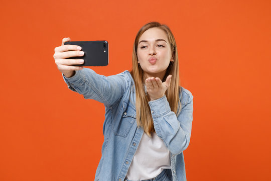 Pretty young woman girl in casual denim clothes posing isolated on orange background in studio. People lifestyle concept. Mock up copy space. Doing selfie shot on mobile phone, blowing send air kiss.
