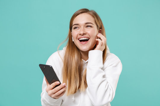 Cheerful Young Woman In Casual White Hoodie Posing Isolated On Blue Turquoise Background Studio Portrait. People Lifestyle Concept. Mock Up Copy Space. Listen Music With Air Pods Using Mobile Phone.