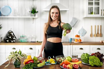 A sporty girl holds .broccoli and stands in the kitchen near a table with fruits and other useful foods. Healthy eating concept