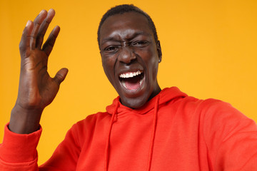 Close up of crazy screaming young african american man guy in red streetwear hoodie posing isolated on yellow wall background in studio. People lifestyle concept. Doing selfie shot on mobile phone.