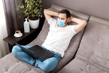Happy relaxed young caucasian man working from home wearing protective mask using laptopinternet. Cozy Home office, workplace on sofa coronavirus pandemic, covid 19 quarantine. Remote work, freelancer