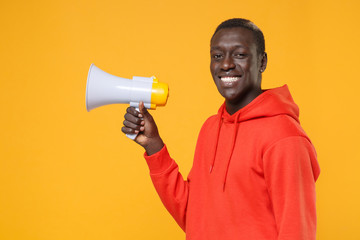 Side view of smiling young african american man guy in red streetwear hoodie posing isolated on yellow wall background. People lifestyle concept. Mock up copy space. Hold megaphone, looking camera.