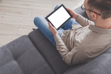 Internet surfing and conference calling. A man uses a tablet computer while sitting on a sofa at home. Dressed in everyday clothes. Tablet with blank screen