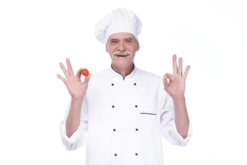 Professional elderly chef in white uniform and hat, holding tomato while staying on white background