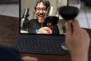 Two middle-aged friends smiling and toasting with red wine during a video call - Connecting and celebrating virtually in the new normal