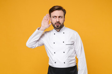 Curious young bearded male chef cook or baker man in white uniform shirt posing isolated on yellow...