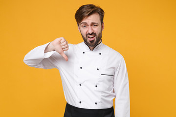 Dissatisfied young bearded male chef cook or baker man in white uniform shirt posing isolated on yellow wall background studio portrait. Cooking food concept. Mock up copy space. Showing thumb down.