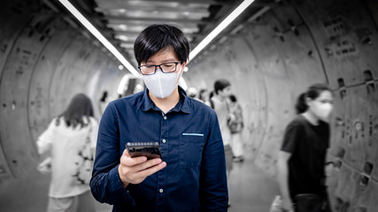 Fototapeta na wymiar Asian man wearing face mask using smartphone in subway tunnel with crowded people walking. Wuhan coronavirus (COVID-19) pandemic protection in public area. Health care and medical concept