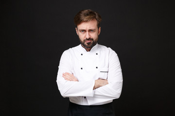 Confused young bearded male chef cook or baker man in white uniform shirt posing isolated on black wall background studio portrait. Cooking food concept. Mock up copy space. Holding hands crossed.