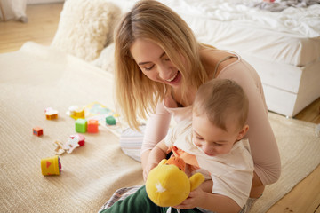 Indoor shot of playful charming little boy sitting on mother's lap holding duck toy and laughing having fun at home. Beautiful young female enjoying social distancing, bonding with her infant son