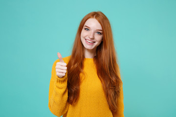 Cheerful young redhead woman girl in yellow sweater posing isolated on blue turquoise wall background studio portrait. People sincere emotions lifestyle concept. Mock up copy space. Showing thumb up.