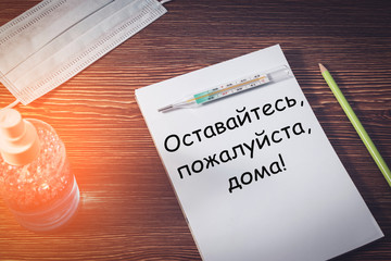 On a wooden table is a Notepad with the inscription "please stay at home", a pencil, detergent, medical mask and thermometer, top view. Address in Russian