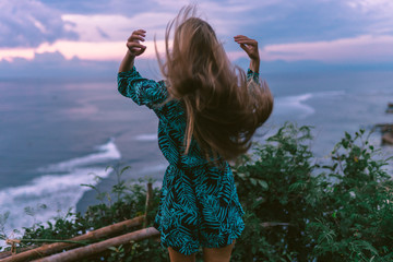 A beautiful girl stands on a cliff overlooking the waves of the blue ocean. Tropical paradise and exotic nature on the island of Bali. Model with different emotions of happiness and joy.