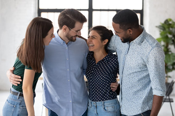 Obraz premium Overjoyed multiethnic colleagues stand hugging at workplace show unity and cooperation, smiling diverse multiracial businesspeople embrace have fun enjoy break in office, teamwork, employment concept