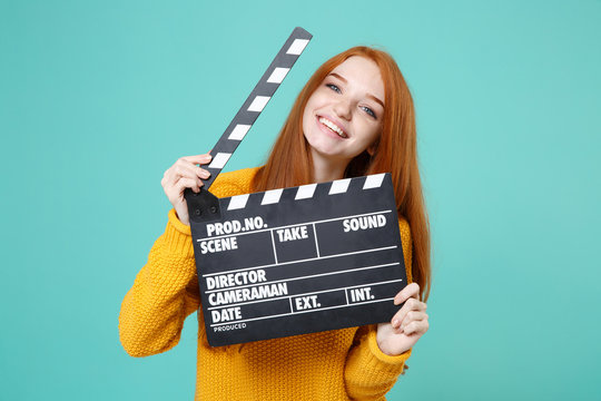 Smiling Young Redhead Woman Girl In Yellow Sweater Posing Isolated On Blue Turquoise Background In Studio. People Lifestyle Concept. Mock Up Copy Space. Hold Classic Black Film Making Clapperboard.