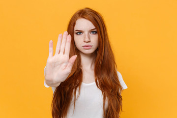 Displeased young redhead woman girl in white blank empty t-shirt posing isolated on yellow background studio portrait. People lifestyle concept. Mock up copy space. Showing stop gesture with palm.
