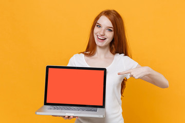 Funny young redhead woman girl in white t-shirt posing isolated on yellow background. People lifestyle concept. Mock up copy space. Pointing index finger on laptop pc computer with blank empty screen.