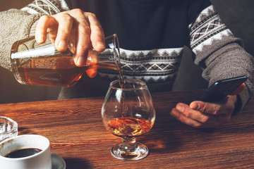 A man pours cognac from a decanter into a glass and holds a smartphone in his hand. There is a Cup...