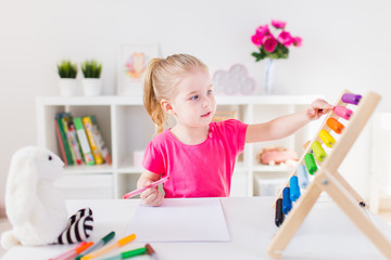 Little smiling blond girl sitting at the white desk and counting on the colourful abacus in the classroom. Home education