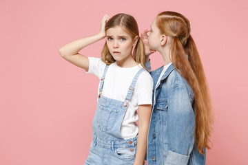 Upset little kids girls 12-13 years old in white t-shirt, denim clothes isolated on pastel pink background. Childhood lifestyle concept. Mock up copy space. Whispering secret behind hand sharing news.