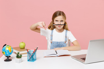 Displeased little kid schoolgirl 12-13 years old study at white desk with pc laptop isolated on pink background. School distance education at home concept. Hold pencil with lips, showing thumb down.