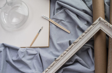 View of a creative space: brushes, vintage frame and blank paper on blue fabric.