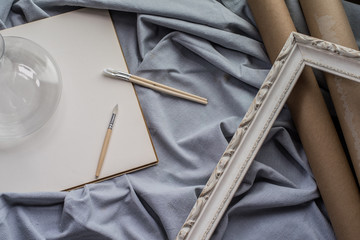 View of a creative space: brushes, vintage frame and blank paper on blue fabric.