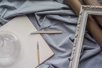 View of a creative space: brushes, vintage frame and blank paper on blue fabric.
